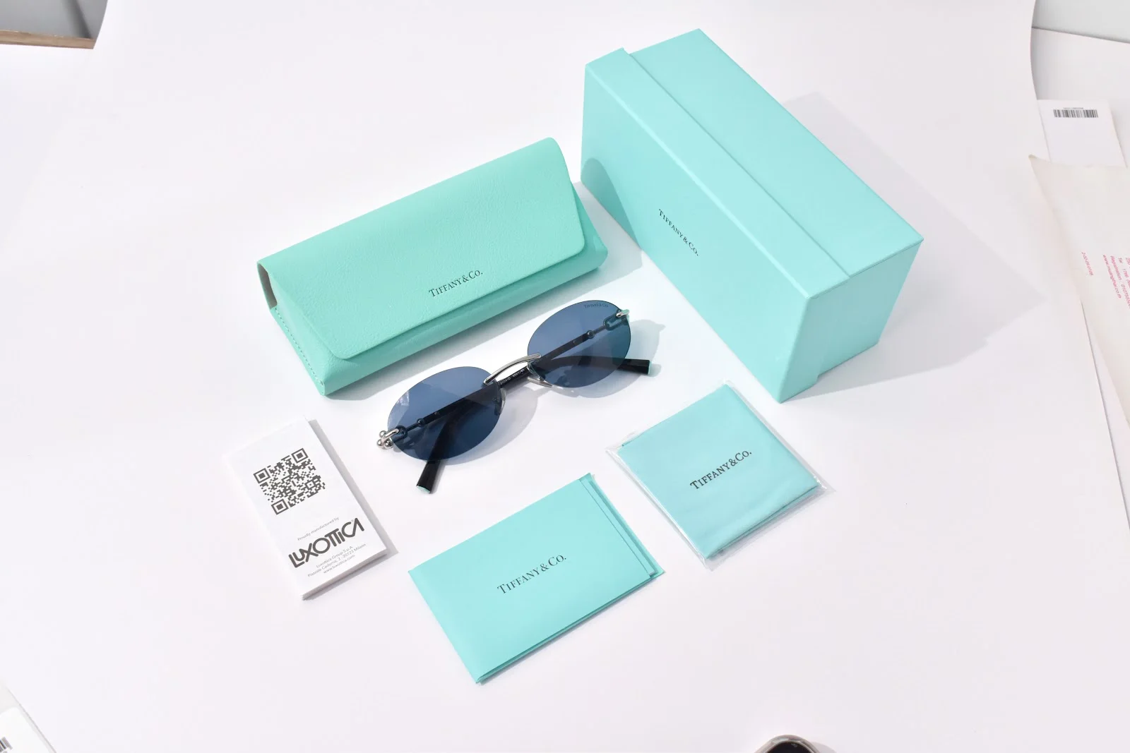กล่องกระดาษสีฟ้า เคสแว่นแข็งสีฟ้า ผ้าเช็ดแว่น และการ์ดคิวอาร์ของ Tiffany & Co. วางคู่แว่นกันแดด TF3104D